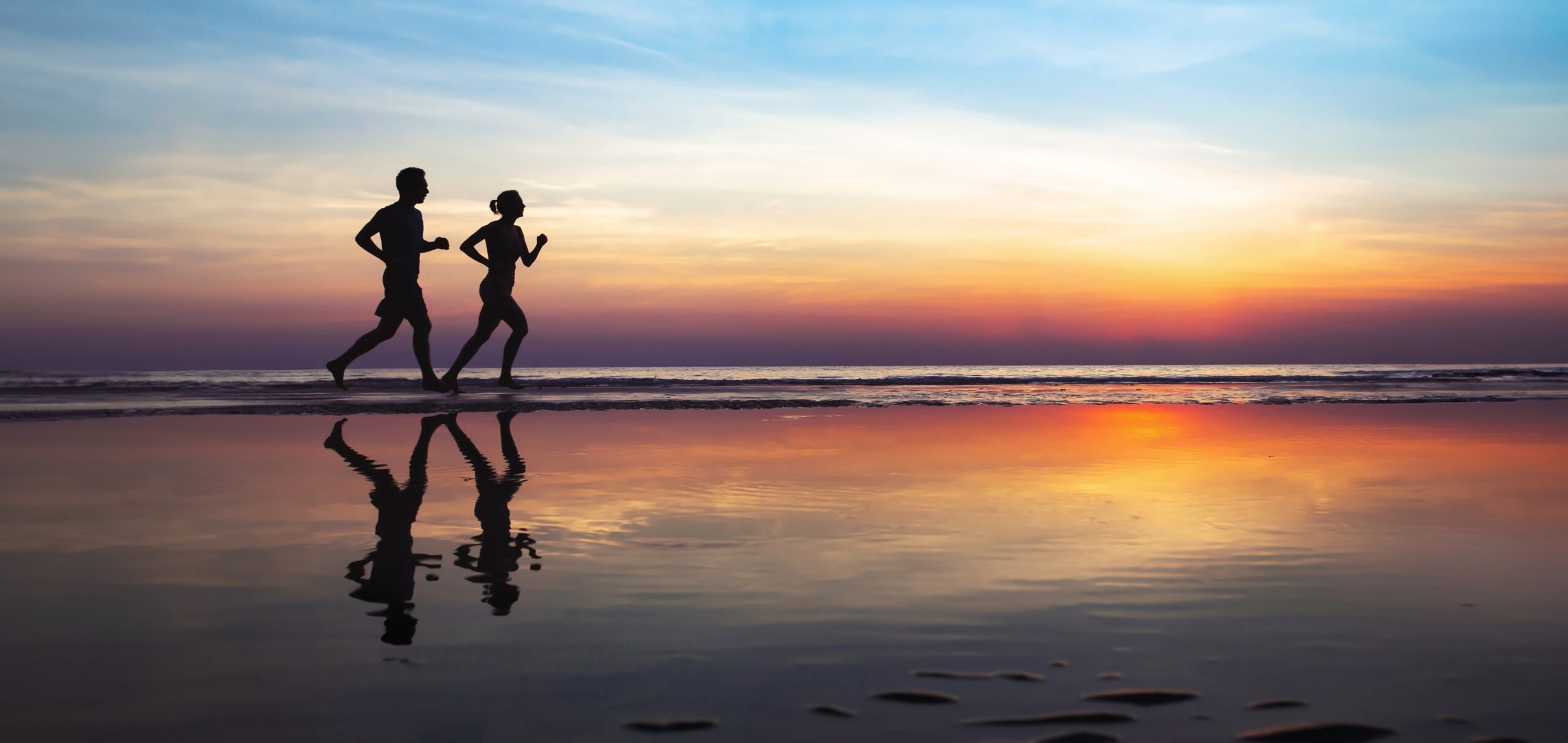 Two runners run on the beach in the evening and reflect in the wet sand together with the sunset
